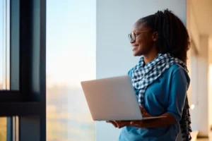 Young Black Woman With Glasses Holding Laptop And Looking Out Window