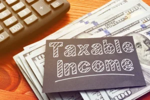 Black Card With White Font "Taxable Income" Written On Top Of Stack Of Cash On Desk