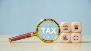 Magnifying Glass Over Word "Tax" Next To Stack Of Wooden Blocks With Graphics On Each